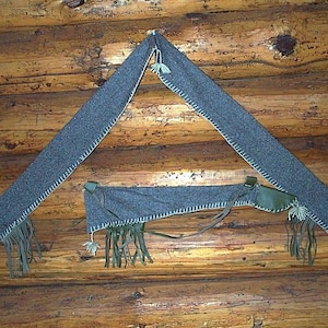 以下が含まれることがあります： 濃い灰色のフェルトのような素材が白い糸で縫い付けられ、木の壁に掛けられています。 このアイテムは、フリンジと革のアクセントが付いた三角形の形をしています。 このアイテムはおそらく装飾品です。