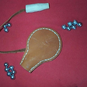 Op de afbeelding: Een bruin leren zakje met witte stiksels, een houten handvat en een leren riem. Verschillende kleine, zilverkleurige metalen ballen liggen verspreid rond het zakje op een rode achtergrond. Dit item is waarschijnlijk een katapult munitiezakje.