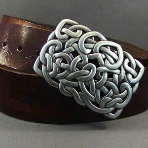 May include: A brown leather belt with a silver Celtic knot buckle. The buckle is intricate and detailed, with a woven design.