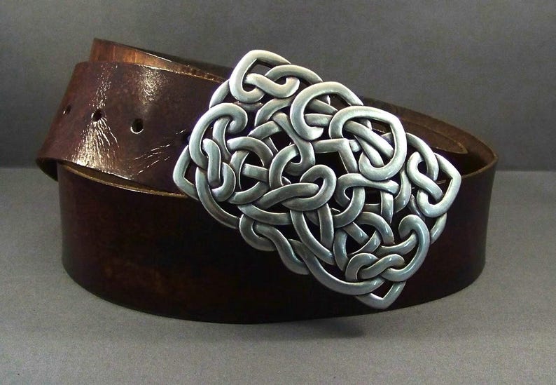 May include: A brown leather belt with a silver Celtic knot buckle. The buckle is intricate and detailed, with a woven design.