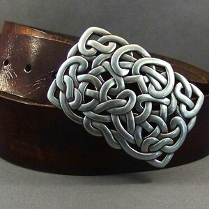 May include: A brown leather belt with a silver Celtic knot buckle. The buckle is intricate and detailed, with a woven design.