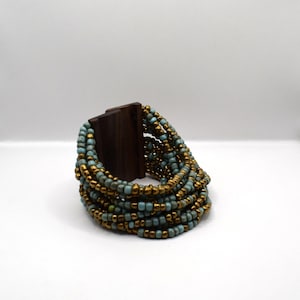 Può includere: Un braccialetto multifilo realizzato con perline turchesi e dorate. Il braccialetto è avvolto attorno a un blocco di legno.