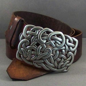 May include: A brown leather belt with a silver Celtic knot buckle. The buckle is intricate and detailed, with a raised design.