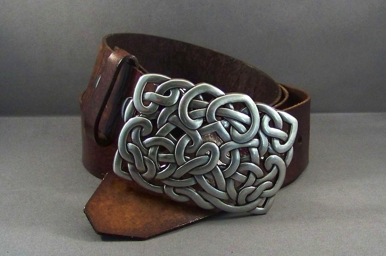May include: A brown leather belt with a silver Celtic knot buckle. The buckle is intricate and detailed, with a raised design.