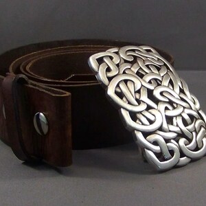 May include: A brown leather belt with a silver Celtic knot buckle. The buckle is intricate and detailed, with a woven pattern.
