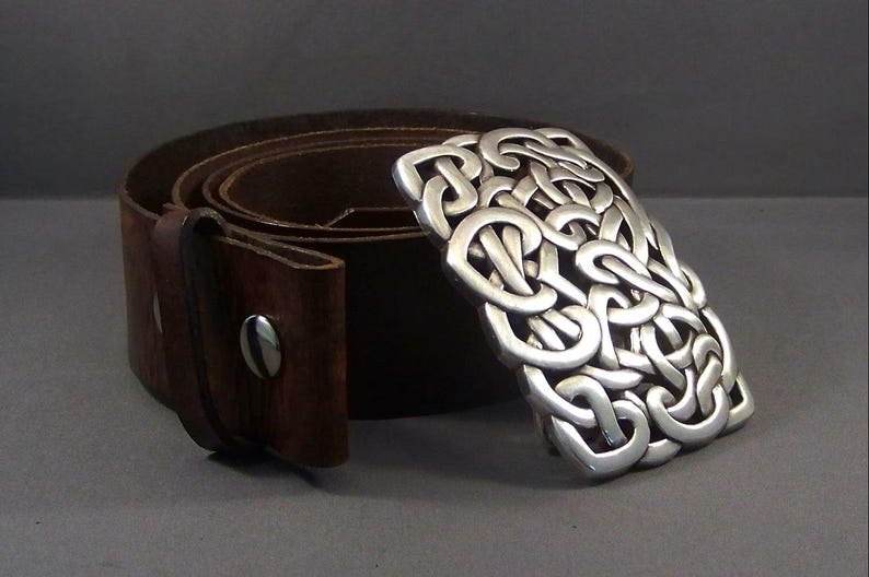 May include: A brown leather belt with a silver Celtic knot buckle. The buckle is intricate and detailed, with a woven pattern.