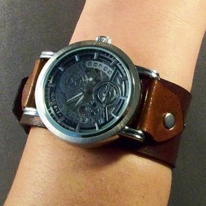 May include: A silver-toned wristwatch with a detailed, dark gray face and a brown leather cuff band. The watch features a complex dial design with visible gears and numerals. The band is secured with a silver button.