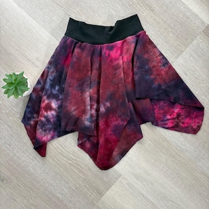 May include: A tie-dye skirt with a black waistband. The skirt is made of a soft, flowing fabric and has a asymmetrical hemline. The colors are a mix of red, purple, and black.