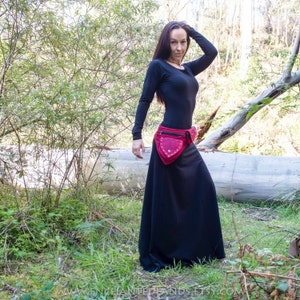 以下が含まれることがあります： 黒い長袖シャツと黒いロングスカートに赤いスタッズのベルトをつけた女性。彼女は倒れた丸太の後ろにある森の中に立っている。