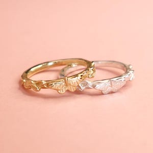 May include: Two rings, one gold and one silver, sit on a pale pink surface. Both rings have a repeating leaf pattern around the band. The gold ring is more detailed. The rings are suitable for everyday wear.