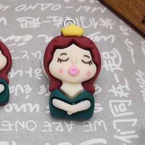 May include: Two green and burgundy polymer clay charms of a princess with a gold crown and closed eyes. The charms have a hole at the top for hanging.