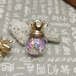 Könnte beinhalten: Zwei goldfarbene Flaschenverschlüsse mit Kronendesign. Jeder Verschluss enthält eine klare Glasflasche, die mit bunten Glitzersternen gefüllt ist.