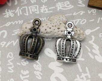 May include: Two small crown charms, one bronze-toned and one silver-toned, sit on a small, light-colored stone. The crowns have intricate details and a loop at the top for attaching to jewelry or other crafts. A red rose and a wooden box are in the background.
