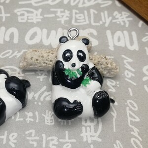 May include: Two panda bear charms. One panda is lying down, and the other is sitting up. Both pandas are black and white with green bamboo. Each charm has a silver loop at the top for hanging.