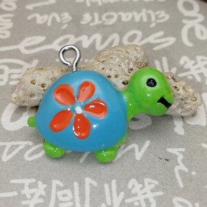 Puede incluir: Un pequeño dije de tortuga de colores brillantes. La tortuga tiene un caparazón azul con un diseño de flor naranja, cabeza y patas verdes. Tiene un lazo plateado en la parte superior para colgarlo.