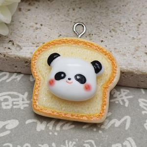 May include: A small, white panda bear charm with black eyes and ears, sitting on a yellow and orange piece of bread. The charm has a silver loop for hanging.