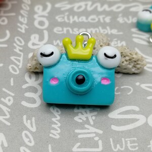 May include: Two blue and yellow camera charms with googly eyes and a crown on one. The charms are made of polymer clay and have a whimsical, cartoonish style.