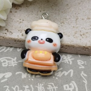 May include: A small, white and black panda bear charm with a chef's hat. The panda is wearing an orange apron with yellow accents. The charm is made of polymer clay and has a silver loop for hanging.