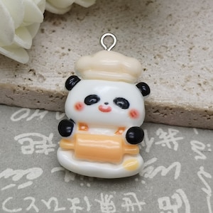 May include: A white panda bear charm with black eyes and a chef's hat, holding a rolling pin. The charm has a silver loop for attaching to a necklace or bracelet.