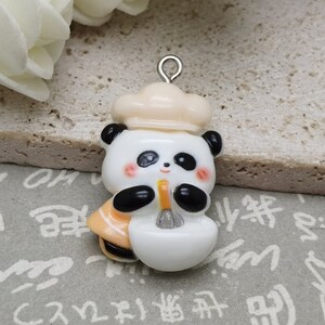May include: A white ceramic panda bear charm with a chef's hat, holding a spoon and a bowl. The charm has black eyes, a black nose, and black paws. It is attached to a silver loop.