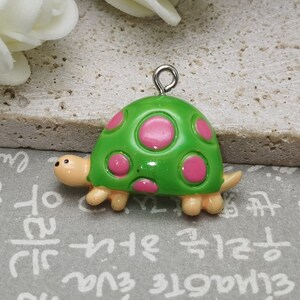 Puede incluir: Un amuleto de tortuga verde con lunares rosas. La tortuga tiene un lazo plateado para colgar.