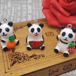 May include: Three white ceramic panda bear charms with black eyes and ears. Each panda is holding a different fruit: a pineapple, a watermelon, and a strawberry.