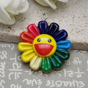 Puede incluir: Un amuleto de flor arcoíris colorido con una cara amarilla sonriente. La flor está hecha de resina y tiene un lazo plateado para sujetarlo a un collar o pulsera.
