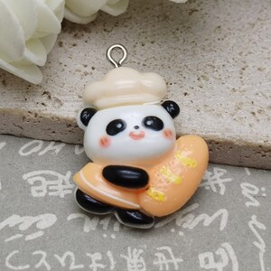 May include: A white panda bear wearing a white chef's hat holds a light orange bread loaf. The panda bear charm has a silver loop for hanging.