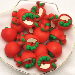 May include: A white bowl with a gold rim filled with red apple-shaped silicone beads. Each bead has a green stem and a green worm inside the apple.