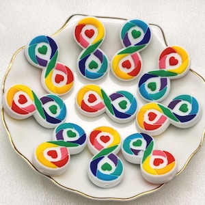 Puede incluir: Un plato blanco con 10 amuletos coloridos en forma de símbolo de infinito. Cada amuleto está hecho de plástico blanco con un diseño de arcoíris y un pequeño corazón en el centro.