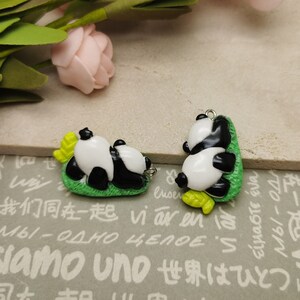 May include: Two black and white panda bear charms with green leaves and yellow accents. The charms are made of polymer clay and have a hole for attaching to a necklace or bracelet.