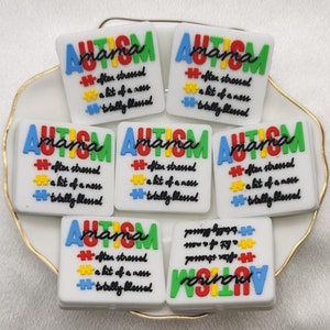 Könnte beinhalten: Sechs weiße quadratische Harz-Anhänger mit den Worten "Autism mama" und einem bunten Puzzle-Motiv. Der Text lautet "oft gestresst, ein bisschen Chaos, total gesegnet".