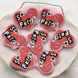 Puede incluir: Ocho dijes en forma de corazón rosa con el texto "So Fetch" en letras negras. Los dijes tienen un diseño rosa y rojo con una impresión de labios.