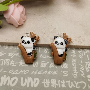 May include: Two brown and white panda bear charms hanging from silver loops. Each panda is sitting on a brown tree branch with green leaves. The charms are on a gray and white patterned surface.