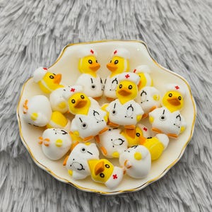 May include: A collection of yellow and white duck figurines, some wearing doctor coats and hats with red crosses, arranged in a white seashell-shaped dish with a gold rim. The ducks have orange beaks and are on a fluffy gray surface.