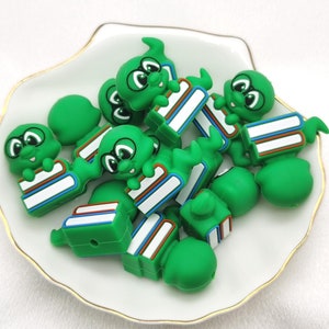 May include: A pile of green silicone bookworm charms with googly eyes and a stack of books. The charms are in a white ceramic dish with a gold rim.
