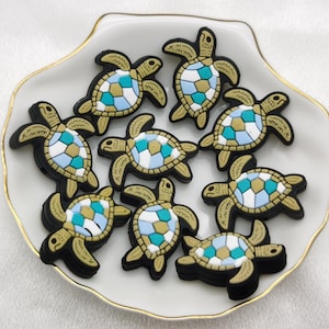 May include: A collection of nine colorful turtle charms with a mosaic design in shades of blue, white, and gold. The charms are arranged on a white plate with a gold rim.