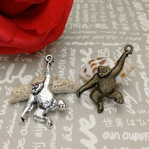 May include: Two silver monkey charms hanging from a loop. The monkey on the left is hanging upside down with its arms raised. The monkey on the right is standing with its arms raised. Both monkeys have a detailed design.