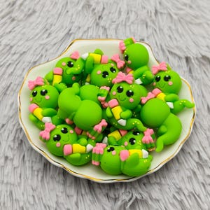 May include: A collection of green, cartoon-like cactus figurines with pink bows and smiling faces, arranged in a decorative, scalloped-edge dish. Each figurine holds a yellow and pink pencil. The dish has a gold trim.