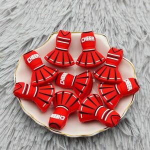 Puede incluir: Un conjunto de pequeños dijes de silicona roja con forma de faldas de animadora, cada uno con la palabra CHEER en letras blancas, líneas negras y rayas horizontales blancas, dispuestos sobre un plato blanco con bordes festoneados.