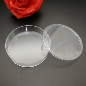 Puede incluir: Un recipiente redondo de plástico transparente con tapa. El recipiente está vacío y la tapa está abierta. El recipiente está sobre una superficie negra.