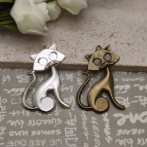 May include: Two metal cat charms. One is silver-toned, the other is bronze-toned. Each charm features a stylized cat silhouette with a head, body, and curled tail. The cat's face has large eyes and a small nose. The charms have a loop at the top for hanging.