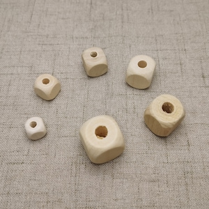 Può includere: Sei perline quadrate in legno con un foro al centro. Le perline sono di dimensioni diverse e sono disposte in cerchio su una superficie di tessuto beige.