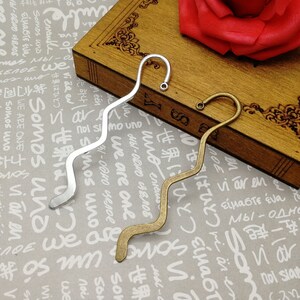 May include: Two metal bookmark designs with a wavy shape and a hook at the top. The bookmark on the left is silver and the bookmark on the right is gold.