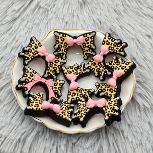 Op de afbeelding: Een verzameling accessoires in de vorm van strikken met luipaardprint en roze strikken, gerangschikt op een wit bord met een gouden rand. De strikken hebben een zwarte rand, tegen een pluizige, grijze achtergrond.