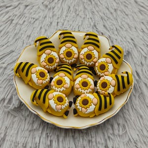 May include: A collection of yellow and black bee-themed gnome figurines with sunflower faces, arranged on a white and gold-rimmed plate. The gnomes have striped hats and are set against a fluffy, gray background.