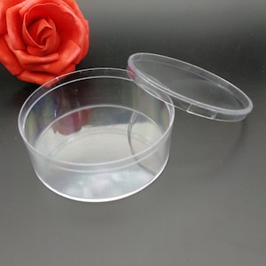 Op de afbeelding: Een ronde, transparante plastic container met een deksel. De container is leeg en het deksel is eraf. De container heeft een diameter van ongeveer 7,5 cm.