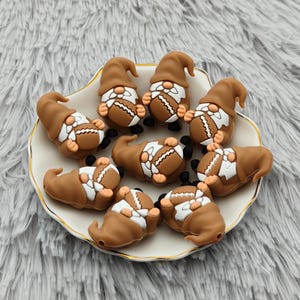 May include: A collection of brown gnome-shaped beads, each holding a football. The gnomes have white beards and hats, and black shoes. They are arranged on a white plate with a gold rim, set against a fluffy, gray background.