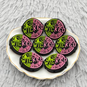 May include: Seven black, round charms with the word "WICKED" in pink and green. They are displayed on a white plate with a gold rim, against a fluffy, white background. The charms also feature the text "I HAVE BEEN CHANGED FOR GOOD".