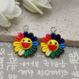 Puede incluir: Dos abalorios de flores arcoíris con una carita sonriente en el centro. Los abalorios son de metal y tienen un acabado plateado.